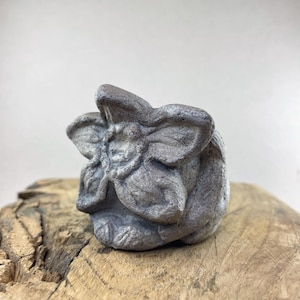 Könnte beinhalten: Kleine, graue Steinskulptur einer stilisierten Blume. Die Skulptur hat detaillierte Blütenblätter und steht auf einem Stück verwittertem Holz. Die Blume ist in einem gedämpften Grauton gehalten, und das Holz hat einen natürlichen, hellbraunen Farbton.