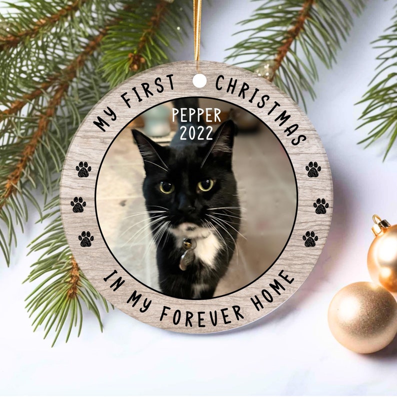 May include: A round wooden ornament with a black and white cat photo in the center. The ornament has the text "My First Christmas" at the top, "Pepper 2022" in the photo, and "In My Forever Home" at the bottom.