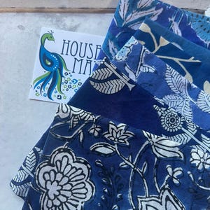 May include: A stack of blue fabric napkins with white floral and leaf patterns. The napkins are in various shades of blue. A card with a peacock illustration and the text "HOUSE MADE" is visible.
