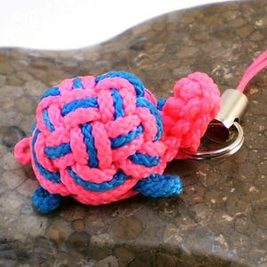 Può includere: Un portachiavi a forma di tartaruga annodata rosa e blu. La tartaruga è realizzata con un cordoncino intrecciato e ha un anello di metallo argentato e un cordino blu.