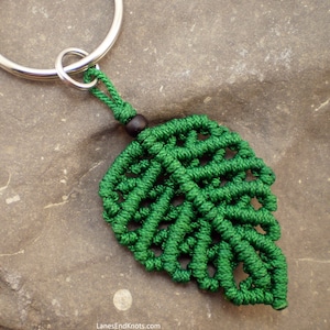 May include: Green leaf-shaped keychain made with macrame knotting technique. The keychain features a silver key ring and a black bead.