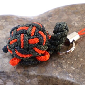 May include: A small, red and green turtle keychain made from braided cord. The turtle is attached to a silver key ring with a black and red cord.