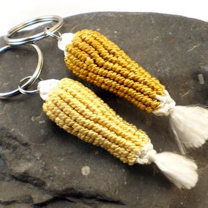 May include: Two corn on the cob keychains, one gold and one yellow, with white tassels. The keychains are attached to silver key rings.