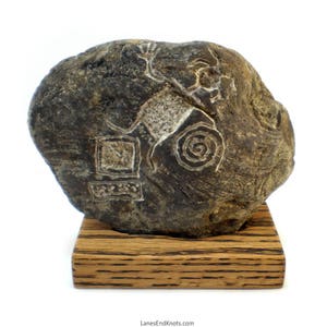 May include: A dark grey stone with a carved image of a person dancing with a spiral design. The stone is displayed on a wooden base.