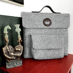 Puede incluir: Maletín gris de fieltro con asa de cuero marrón y emblema de corona. El maletín tiene cierre de solapa y un bolsillo delantero más pequeño. Una escultura de bronce de dos figuras con cabezas de piezas de ajedrez está sobre una mesa roja.