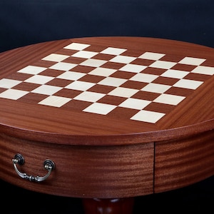 Peut inclure: Table de jeu d'échecs ronde en bois brun foncé. Le plateau présente un motif de damier avec des cases claires et foncées. Un petit tiroir avec une poignée métallique est visible.