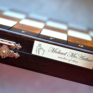 Può includere: Una scacchiera di legno con una placca in ottone che recita "Michael McArthur, maestro di scacchi". La scacchiera ha una finitura marrone scuro e una chiusura in ottone.