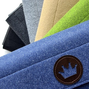 Può includere: Un mucchio di pochette in feltro di vari colori, tra cui nero, grigio, beige, verde e blu. La pochette blu ha una toppa in pelle marrone con un disegno a corona.