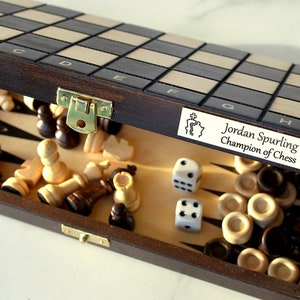 Può includere: Un set da gioco di scacchi e backgammon in legno con un esterno marrone e una scacchiera bianca e nera all'interno. Il set include pezzi degli scacchi, pezzi di backgammon e due dadi. Il coperchio della scatola ha una targhetta personalizzata che recita "Jordan Spurling Champion of Chess".