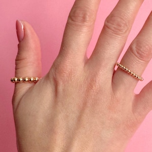 May include: A hand with two gold beaded rings on a pink background. The rings are made of small, round beads strung together. The nails are painted with a light pink nail polish.