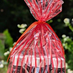 May include: A gift bag filled with small jars of infused honey. The jars are wrapped in a clear cellophane bag with red stripes and tied with a red ribbon. The labels on the jars read "Jalapeno", "Serrano", and "Peach".