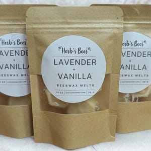Puede incluir: Tres bolsas de papel marrón con ventanas transparentes, cada una etiquetada "Herb's Bees LAVENDER + VANILLA BEESWAX MELTS". Ramas de lavanda moradas en la parte inferior. Las bolsas contienen ceras perfumadas.