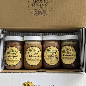 May include: A cardboard box with a sample pack of four small glass jars of whipped honey. The jars are labeled "Herb's Honey Whipped" with different flavors: "La Chocolate", "Spice Cake", "Elderberry", and "Cinnamon Bun". The box has a sticker with the same logo and the text "Whipped Honey Sample Pack".