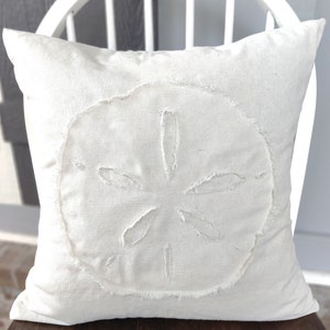 May include: A white throw pillow with a sand dollar design. The sand dollar is outlined with a frayed edge.