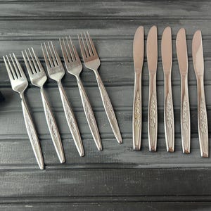 Può includere: Un set di forchette e coltelli in acciaio inossidabile disposti su una superficie di legno grigio scuro. Le forchette hanno quattro rebbi e un motivo floreale decorativo sui manici. I coltelli hanno un design simile.