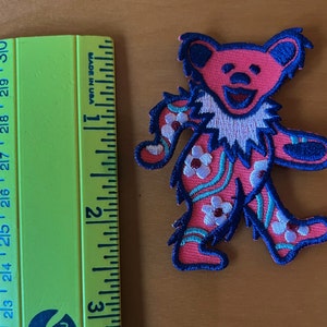 May include: A pink embroidered bear patch with blue accents and white flower details. The patch is next to a green ruler showing inches. The bear has a smiling face and is in a walking pose.