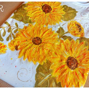 May include: White t-shirt with a hand-painted design of sunflowers in shades of yellow, orange, and green. The sunflowers are arranged in a cluster with leaves and stems.