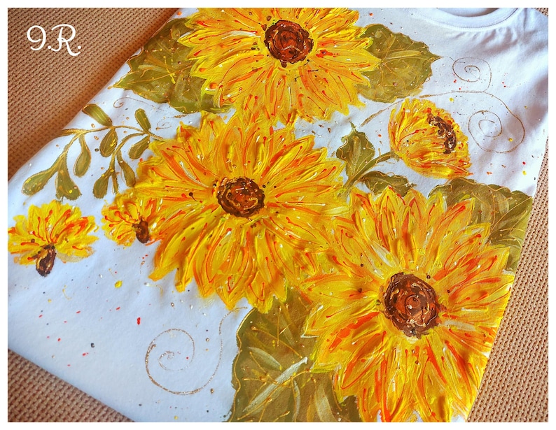 May include: White t-shirt with a hand-painted design of sunflowers in shades of yellow, orange, and green. The sunflowers are arranged in a cluster with leaves and stems.