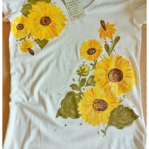 May include: White t-shirt with a hand-painted design of sunflowers and leaves. The sunflowers are yellow with brown centers and the leaves are green. The shirt has a V-neck and short sleeves.