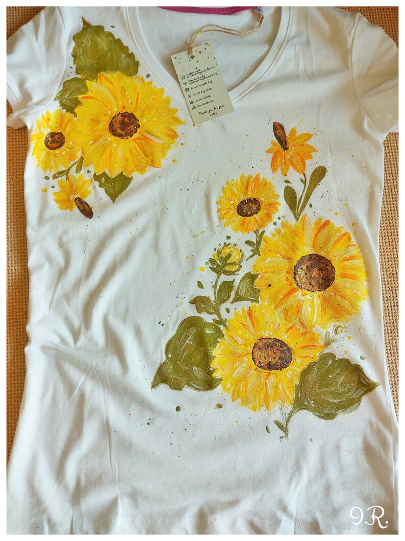 May include: White t-shirt with a hand-painted design of sunflowers and leaves. The sunflowers are yellow with brown centers and the leaves are green. The shirt has a V-neck and short sleeves.