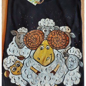 May include: A black t-shirt with a hand-painted design of a cartoon sheep with large horns and four smaller sheep surrounding it. The sheep are all white with black eyes and noses.