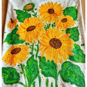 May include: A white t-shirt with a hand-painted design of sunflowers with green leaves. The sunflowers are yellow with brown centers. The design is on the front of the shirt.
