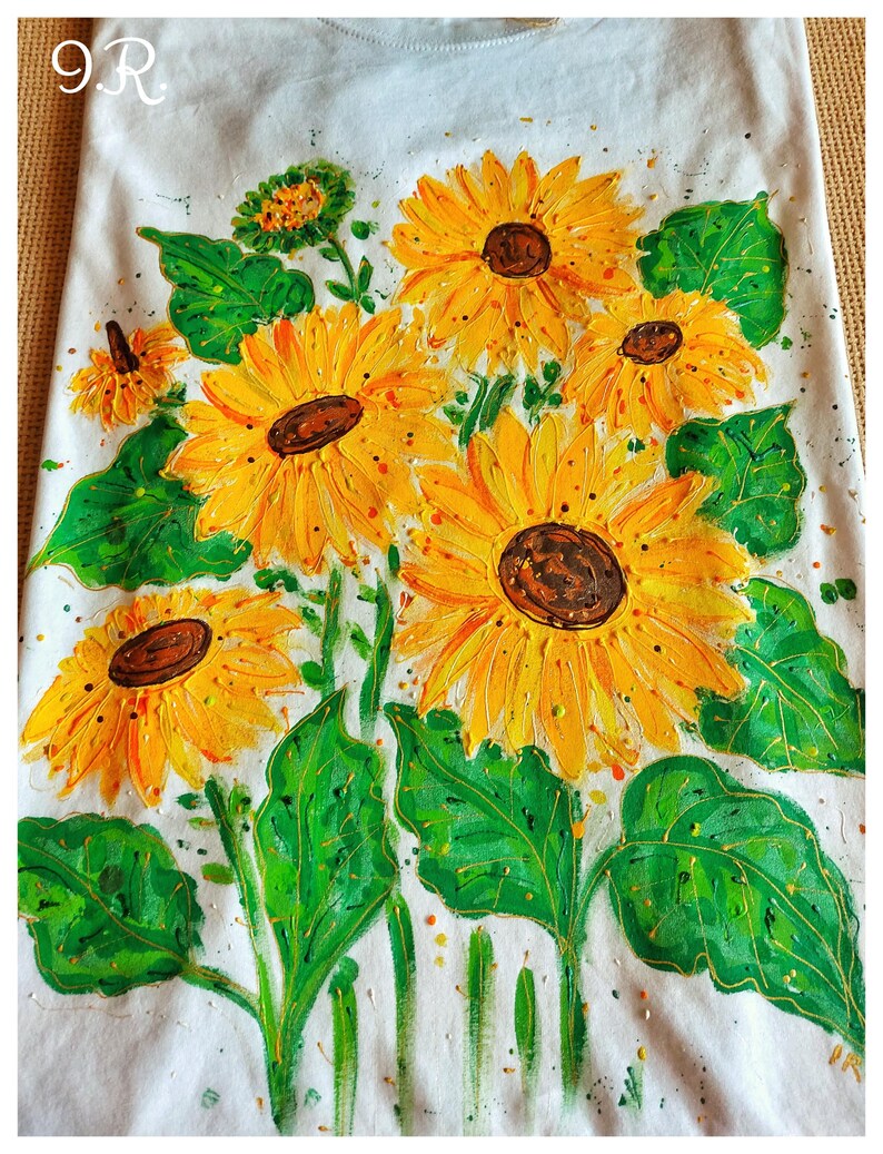 May include: A white t-shirt with a hand-painted design of sunflowers with green leaves. The sunflowers are yellow with brown centers. The design is on the front of the shirt.