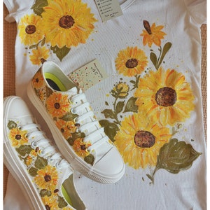 May include: A white t-shirt with a hand-painted sunflower design. The shirt is paired with a pair of white canvas shoes, also hand-painted with sunflowers. The shoes have green laces and a white sole.