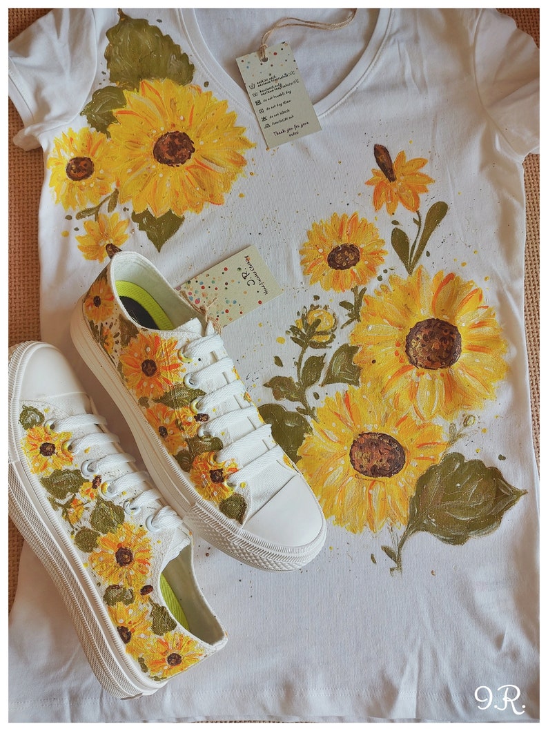 May include: A white t-shirt with a hand-painted sunflower design. The shirt is paired with a pair of white canvas shoes, also hand-painted with sunflowers. The shoes have green laces and a white sole.