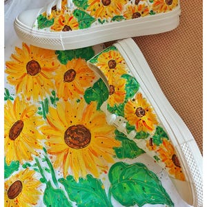 May include: A pair of white canvas shoes hand-painted with a sunflower design. The shoes are on a white surface with a sunflower pattern.