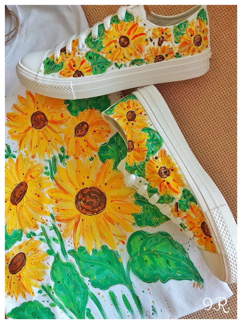 May include: A pair of white canvas shoes hand-painted with a sunflower design. The shoes are on a white surface with a sunflower pattern.