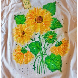 May include: White t-shirt with a hand-painted design of a bouquet of sunflowers with green leaves. The sunflowers are yellow with brown centers. The leaves are green with a light green outline. There are small yellow and orange paint splatters on the shirt.