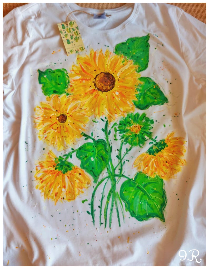 May include: White t-shirt with a hand-painted design of a bouquet of sunflowers with green leaves. The sunflowers are yellow with brown centers. The leaves are green with a light green outline. There are small yellow and orange paint splatters on the shirt.
