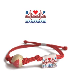 May include: Red and white bracelet with a red heart and the letters "SF" on a white tag. The tag also features a red outline of the Golden Gate Bridge with blue water underneath. The bracelet has a red cord and a red and white bead.