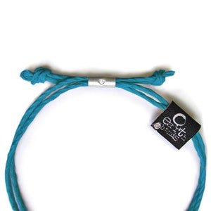 May include: A turquoise blue braided cord bracelet with a silver heart charm and a black tag that reads "Orit's Earth Beads".