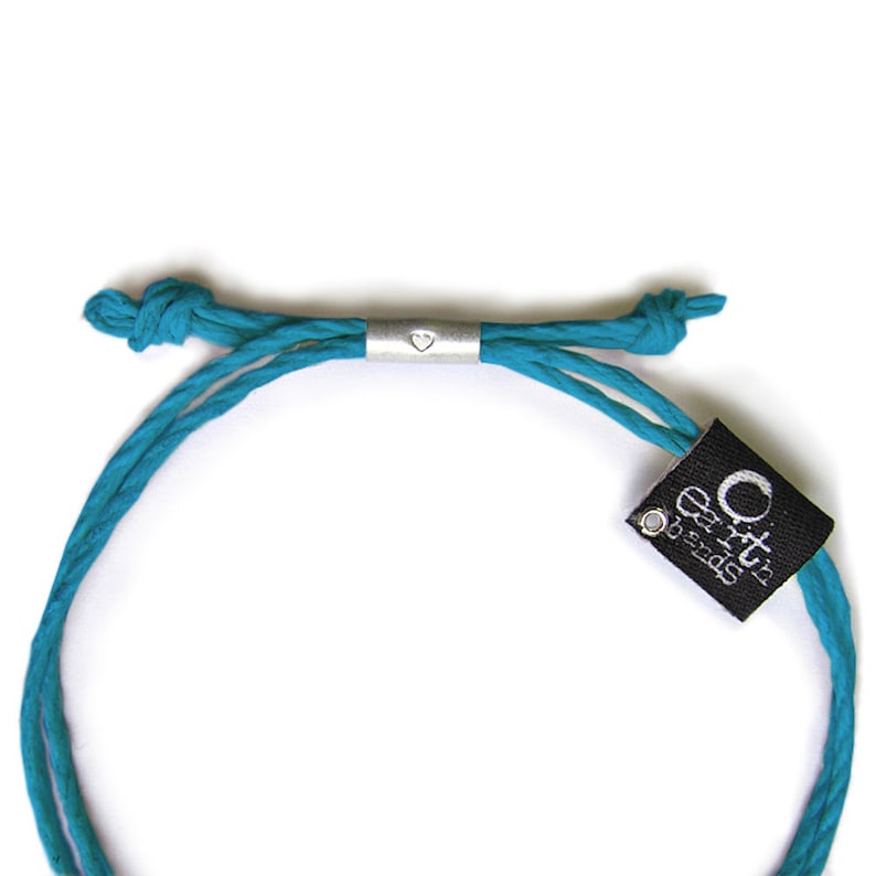 May include: A turquoise blue braided cord bracelet with a silver heart charm and a black tag that reads "Orit's Earth Beads".