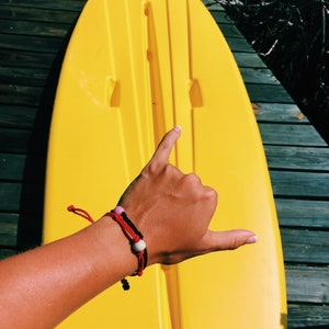 May include: A bright yellow kayak with a person's hand making a shaka sign. The hand is wearing a red and black bracelet with white and pink beads.