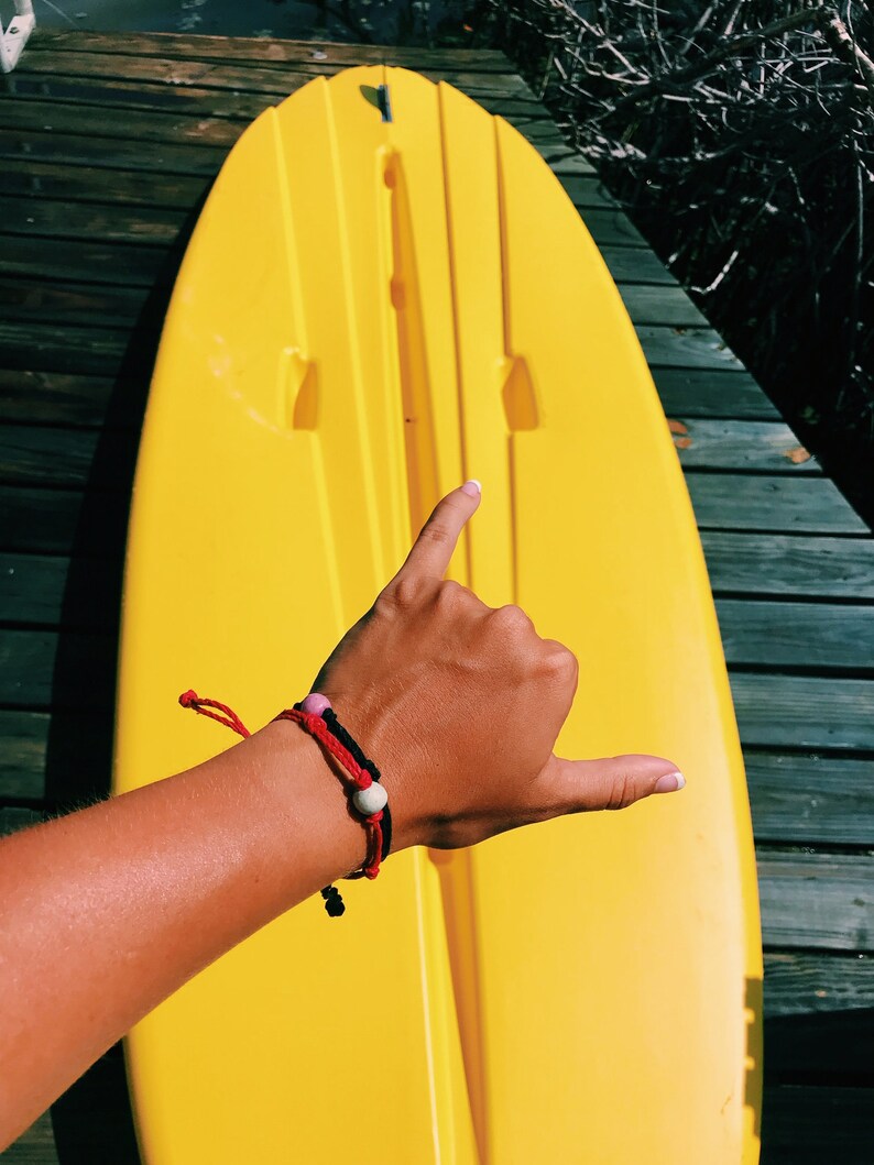 May include: A bright yellow kayak with a person's hand making a shaka sign. The hand is wearing a red and black bracelet with white and pink beads.