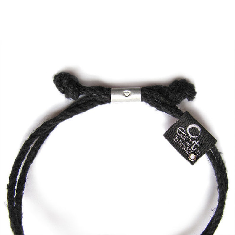 May include: Black cord bracelet with a silver heart charm and a black tag with the text "Orto Designs" in white.