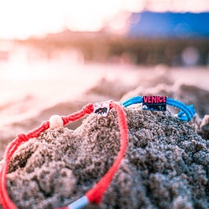 May include: Two colorful friendship bracelets with white beads, one red and one blue, are partially buried in sand. The blue bracelet has a tag that reads "VENICE CALIFORNIA".