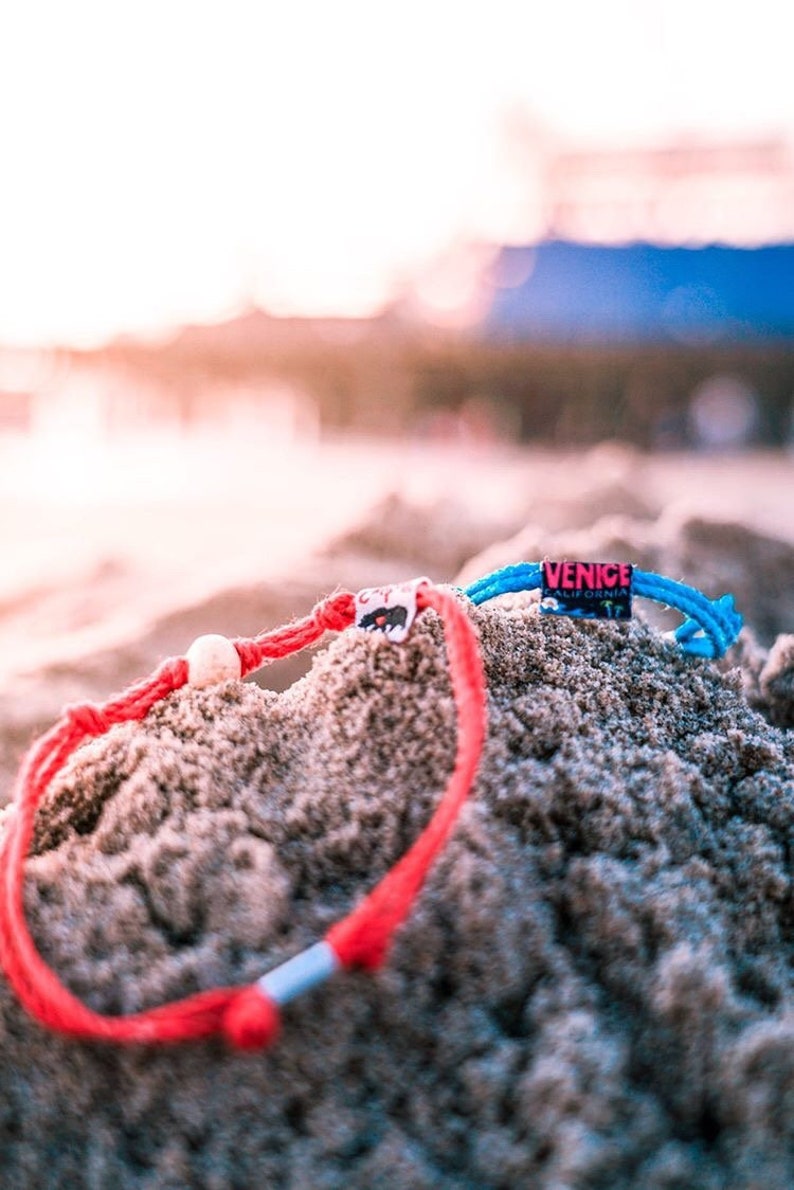 May include: Two colorful friendship bracelets with white beads, one red and one blue, are partially buried in sand. The blue bracelet has a tag that reads "VENICE CALIFORNIA".