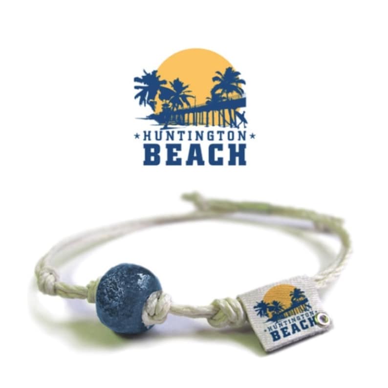 May include: White cord bracelet with a blue glass bead and a small square tag with the text "*HUNTINGTON* BEACH" in blue and yellow.