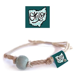 Puede incluir: Un brazalete de cuerda marrón con una cuenta azul claro y una etiqueta turquesa con la palabra "Ohio" y un contorno estilizado del estado de Ohio.