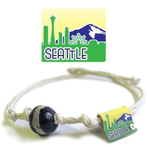 May include: A white cord bracelet with a black and white bead and a green and yellow charm with a silhouette of the Seattle skyline and the word "SEATTLE" printed on it.