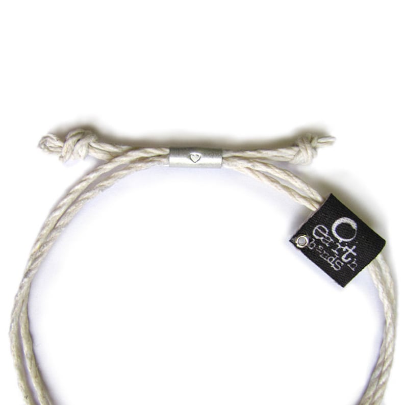 May include: A white braided cord bracelet with a silver heart charm and a black tag that reads "Earth's Treasures".