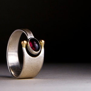 Può includere: Un anello d'argento con finitura spazzolata, con una pietra preziosa rosso intenso incastonata in una lunetta d'oro. L'anello ha un design unico e moderno con accenti dorati su entrambi i lati della gemma.