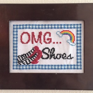 May include: A framed cross-stitch with a blue and white gingham border. The cross-stitch features a pair of red shoes with black and white stripes, a rainbow, and the text "OMG...Shoes".