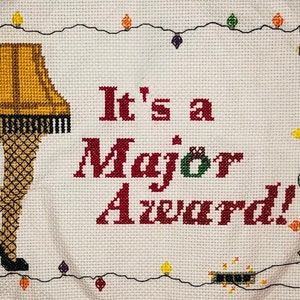 May include: Cross-stitch artwork featuring a leg lamp and the phrase "It's a Major Award!" in red letters. The lamp is yellow and brown, with a black fringe. The text is surrounded by a string of colorful Christmas lights.