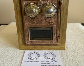 May include: A wooden box with a copper-colored front panel, featuring two circular dials and a small rectangular window. The box has a hinged door and is framed in light-colored wood. A card with instructions for opening the box is in front.