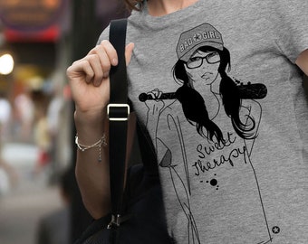 May include: Gray t-shirt with a black and white illustration of a woman wearing a baseball cap that says "Bad Girl" and holding a baseball bat. The text "Sweet Therapy" is printed on the shirt.